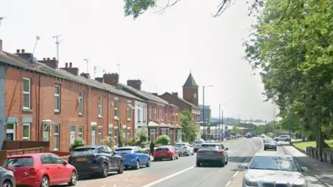 Parked cars and red-bricked terraced houses are on the left hand side of the road with a grassed area and trees on the right hand side.