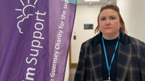 Jenny Murphy pictured with short red hair and hoop earrings. she is wearing a navy and red tartan jacket with a lanyard. Jenny is standing next to a purple banner which says 'Victim Support, the Guernsey charity for the support' on.