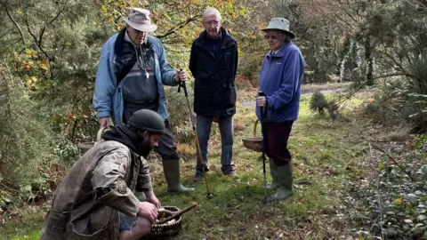 In a clearing in a wood, three people stand around a man giving information about a mushroom he has found. He is crouching down. The others are listening intently. They are all wearing outdoor clothing and carrying baskets. 