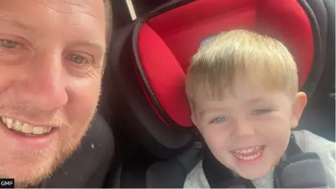 Dylan, with short blond hair, is sitting in a red car seat and smiling. Alongside him is his father Gary, also smiling.