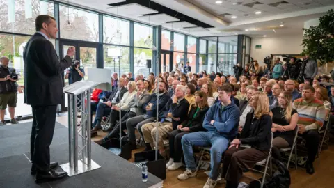 Matthew Horwood Rhun ap Iorwerth speaks from a stage to an audience in a building that has a glass wall on the left hand side. There is a plant visible at the back of the crowd.