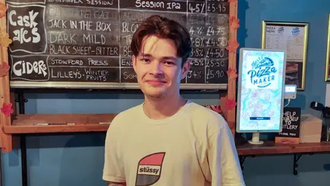 Bobby Bruen with brown hair wearing a white t-shirt with a red and green stussy logo standing in a pub in front of a blackboard featuring drinks on offer. He is smiling.