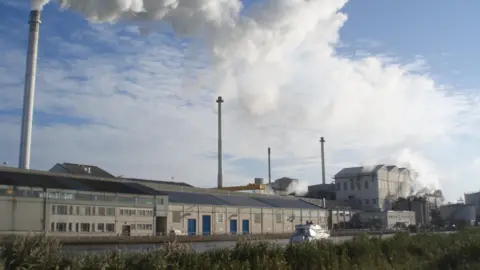 A factory with clouds of steam rising into the air on a sunny day.