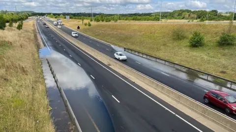 A dual carriageway which is flooded on the nearside lanes on both the east and westbound sections. There is traffic on the road which has trees and grass on either side.