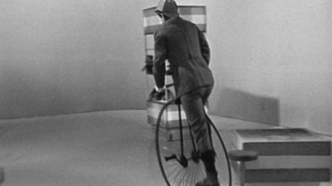 Black and white image of John Noakes on a penny-farthing, cycling away from the camera. He appears to be in riding gear and cycling hat.