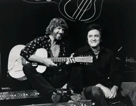 Getty Images Kris Kristofferson poses with a guitar in hand with country music legend Johnny Cash on the set of a TV show in the 1960s.