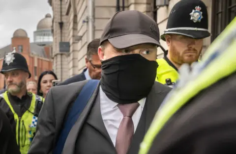 PA Media A man wearing a grey suit, white shirt, tie, black cap and face mask, is seen leaving a court building surrounded by police officers wearing hi-vis vests and black uniforms.