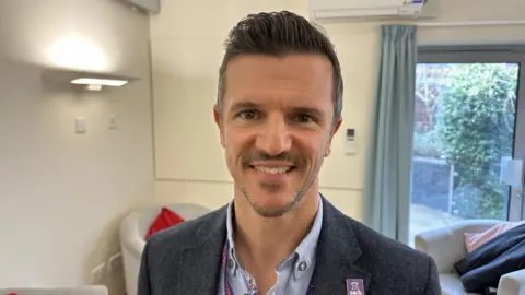 Ben Parker/BBC Kevin Clements wearing a grey blazer with a blue shirt. He has short dark hair.