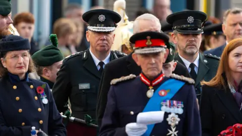 Pacemaker Press A crowd of people are standing close together. The Chief Constable is wearing his PSNI uniform with lapels on his shoulders. Others are wearing army uniforms. A woman is wearing a navy coat with gold buttons and a poppy.
