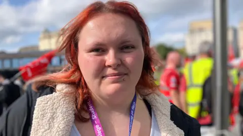 A woman with dyed red hair is standing in a crowd of people. She is seen from her shoulders up. She is looking at the camera.