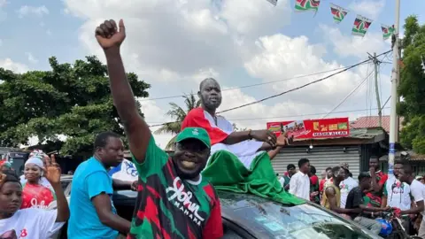 Usman Minjibir / BBC Supporters of John Mahama celebrating on the streets of Accra. One man is sitting on a car, draped in a flag of his party's colours, while another has his fist raised.