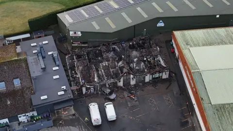 An ariel photo of the buildings destroyed by the fire. The roof has been completely destroyed. A silver car which is also completely burned is right in front of the entrance to one of the buildigs.