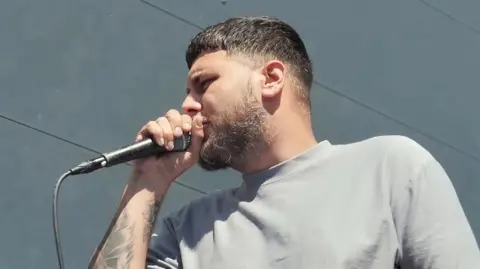 Bearded man with a grey t-shirt singing into a microphone. He has tattoos on his arm and is photographed side-on