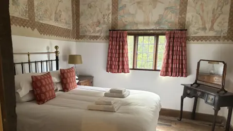 A white bedroom with red patterned curtains and historic-looking paintings across the upper part of the walls. There is a dark wood dressing table in the corner.