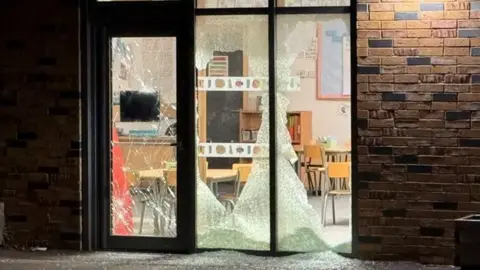 Smashed windows and a door can be seen with glass scattered over the floor outside a classroom at one of the schools.