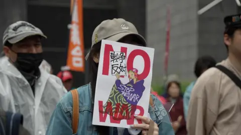 A person holds a sign over their face with the words 'NO WAR'