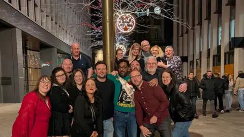 Krissie Edwards A group of men and women standing outside in a group. Behind them are some buildings and they are smiling at the camera. 