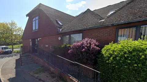 The exterior of the Little Common Surgery in Cooden Sea Road in Bexhill. It is a red-brick building with a brown door, from which there are both steps and an access ramp. Two green and one pink shrub run parallel to the access ramp. 