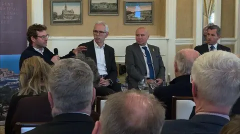 From left to right is Deputy Carl Meerveld, Mat Desforges, Huub den Rooijen and Dr Mark Leybourne sat on a panel. There are people sat in front of them, with their backs to the camera. There are three picture framed images on the dark beige wall behind the panel. 