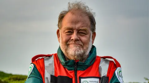 BASICS Devon Dr David Hillebrandt, looking directly at the camera, is wearing a red hi-vis jacket and green jacket. He has a name badge which says "Volunteer" on it. The sky behind him is grey.