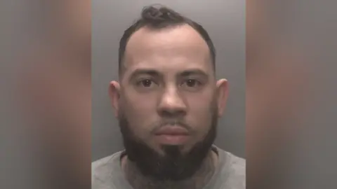 Leicestershire Police A man with dark hair and a beard and brown eyes, wearing a grey top