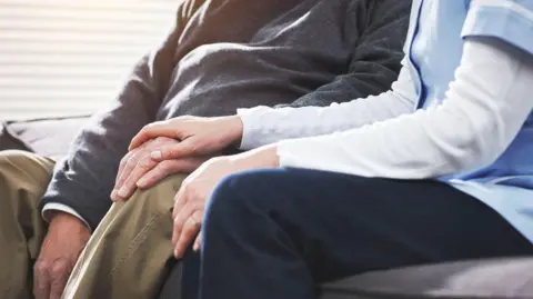 Getty Images Close up of a care workers hand on top of an elderly man's hand. The care worker is wearing a light blue tunic and the man is wearing brown trousers and a grey jumper. Their faces are not shown.
