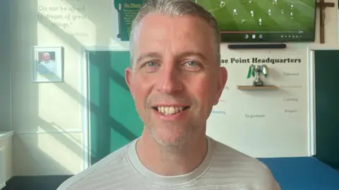 Paddy McMahon, head of St Patrick's RC Primary School, who has short grey haor and a grey top stands in a room in the school with football on a TV screen behind him.