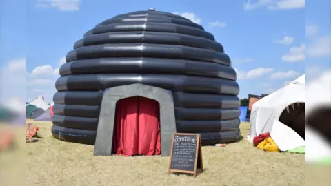 A large black inflatable planetarium is positioned in a field next to other tents and marquees. It has a red curtain covering its entrance. Out the front is a chalk A-board which says Cosmodrome and lists the times of such events as "the dark side of the moon".