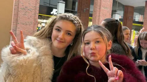 Maisie has long blonde hair with a hairband in, she is wearing a fur jacket and black top. Hannah has brown and blonde hair which is tied back and she is wearing a red fluffy coat.