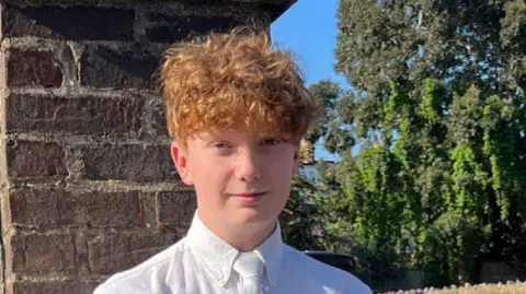 Harry Pitman, a 16 year old boy with curly red hair, standing by a wall on a sunny day