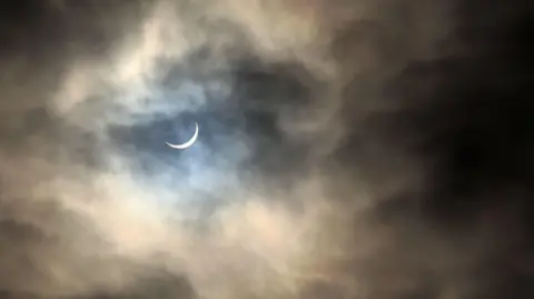 Getty Images A partial solar eclipse in a cloudy night sky. A crescent of white moon can be seen. 