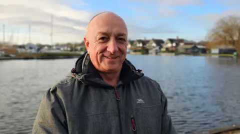 Paul Moseley/BBC Jason Masser is smiling and wearing a grey outdoor coat. Behind him is the River Bure.