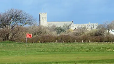 Des Blenkinsopp / Geograph A telephoto shot of Christchurch Priory in the distance, with a putting green and an orange flag of Solent Meads gold course in the foreground. The priory church is a huge stone medieval building with a square tower at one end.