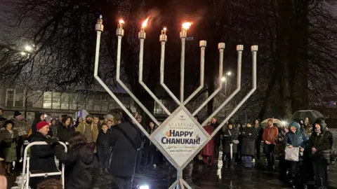 BBC A giant Menorah is lit in Plymouth City Centre. People stand behind it in the dark looking on.