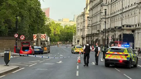 A police car and two policemen are seen in front of a cordon on Grosvenor Place. A car boot is seen open, while debris is seen in the road 
