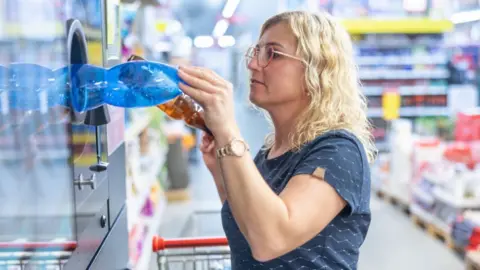 Getty Images A woman with glasses, blonde hair and a blue T-shirt puts plastic bottles into a reverse vending machine in a supermarket. 