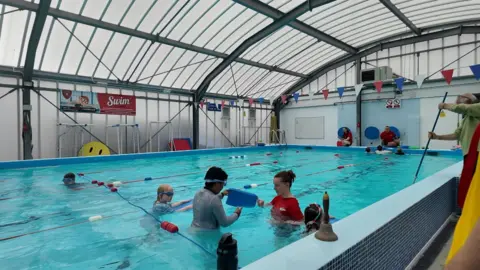 a swimming lesson in progress in a raised swimming pool. The pool is segregated into swimming lanes with a student being shown how to use a float by an instructor