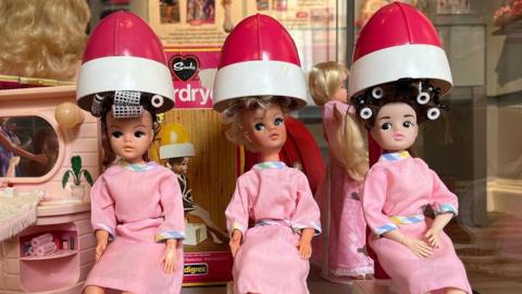 Three dolls sitting in a row wearing pink and having their hair done