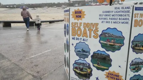 BBC A brightly-coloured advertising board promotes self-drive boats on St Ives seafront. In the background, a woman and a child are walking hand-in-hand toward a beach.