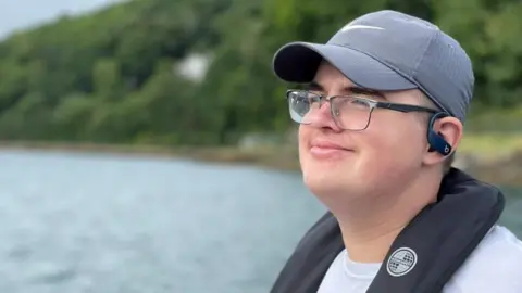 A young man wearing a white T-shirt, black baseball cap, glasses and life jacket is sat on a canoe and is looking out onto the water. He has headphones in his ears