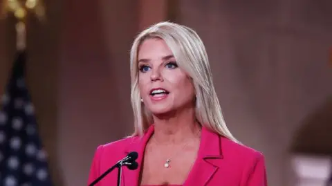 Pam Bondi speaks at the Republican National Convention in 2020. She is standing in front of a lectern with the words "Trump-Pence" on it. She has straight blonde hair and is wearing a pink blazer.