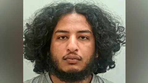 Qusai Abdalla's mugshot. He has a dark beard, brown eyes and shoulder-length, bushy dark hair with a central parting. He is wearing a grey t-shirt and is in front of a plain grey background