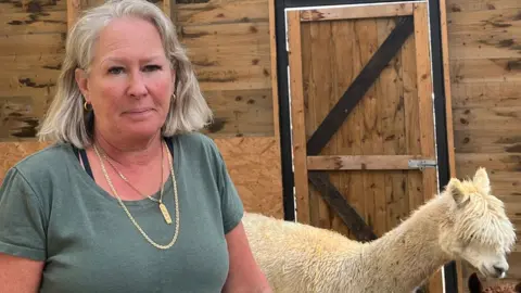 Katherine Ganczakowski/BBC Dawn French with an alpaca. She has shoulder-length grey hair and is wearing a green T-shirt. Behind her is a white alpaca and they are in a wooden barn