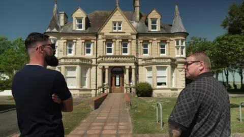 Dan Stanaway and David Bishop stand in front of the grand Victorian school building. The three-storey, stone, double-fronted building has a portico entrance with bay windows either side and small pointed turrets on the corners of the roof.