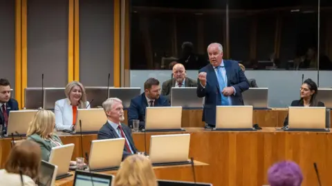 Senedd Cymru Andrew RT Davies speaking in the Senedd, stood up and gesticulating with his right hand, surrounded by seated members of his Conservative group, mostly with neutral expressions with the exception of Laura Anne Jones, who is smiling. All are sat with their computer screens in front of them.