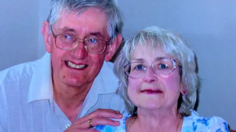 Ray Turnbull Ray is holding on to his wife Jean with his hands on her shoulder and around her back. He is wearing a white shirt, glasses and a wedding ring. They both have grey hair. Ray is looking sideways and Jean, who is wearing a blue and white floral top and glasses, is looking straight at the camera. She has large blue eyes. 