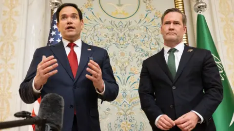 Mark Rubio and Mike Waltz speaking to the media, with flags behind them. 