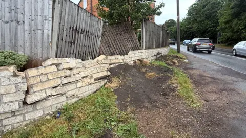 A brick wall and wooden fencing have been crashed into. The wall has several large cracks within the brickwork and has become unstable in places. A large pile of dirt has been pushed towards the brick wall. Wooden fences behind it are dislodged and displaced.