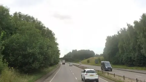 The A50 eastbound in Derbyshire, with a white car at the front of the image and trees either side of the road
