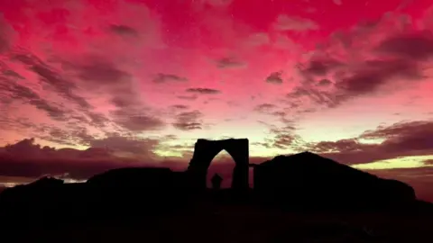 BBC Grosnez Castle in dark lighting with a pink and yellow sky above it and a person stood in the archway.